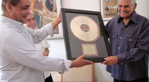Dos hombres sonriendo, uno recibe disco de oro
