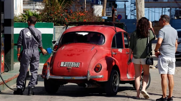 Coche rojo antiguo repostando en gasolinera, personas caminando.