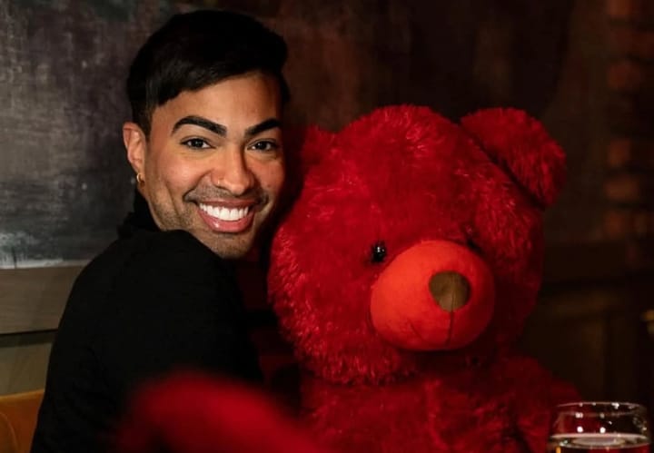 Persona sonriente junto a oso de peluche rojo.